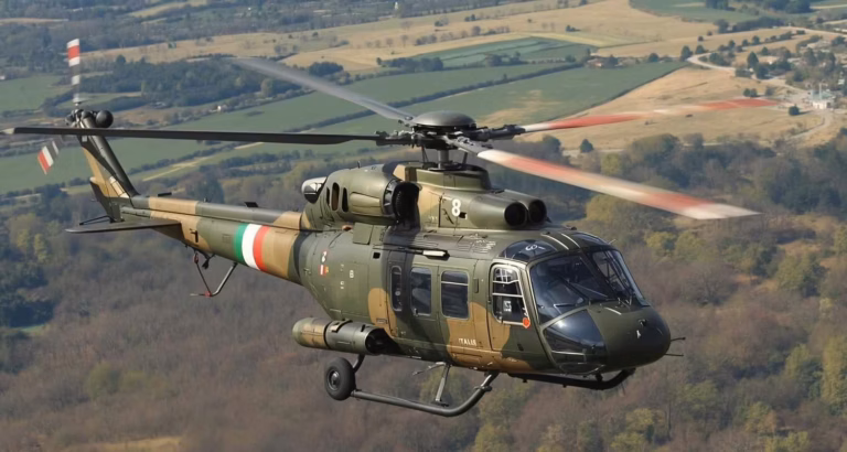 Elicottero militare mimetico in volo sopra un paesaggio collinare, con rotori in movimento e carrello retratto.