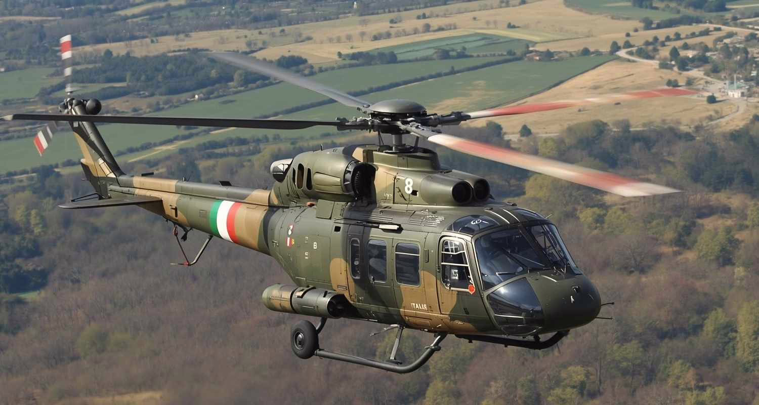 Elicottero militare mimetico in volo sopra un paesaggio collinare, con rotori in movimento e carrello retratto.