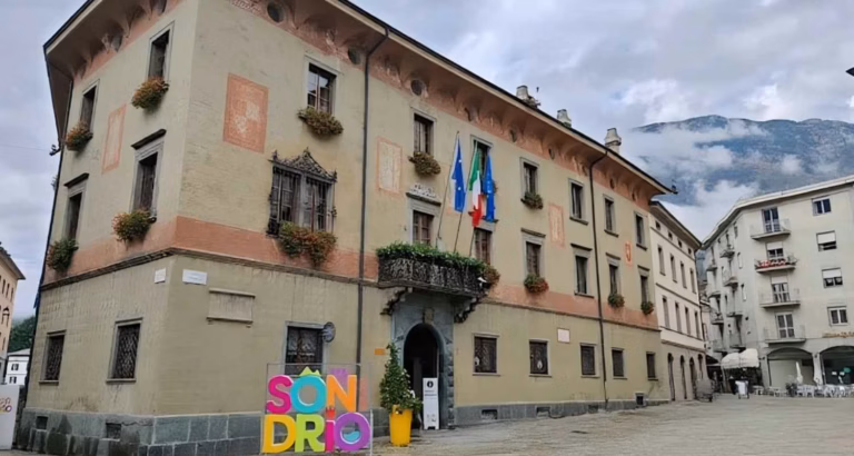Palazzo del Comune di Sondrio con bandiere italiane ed europee e la scritta colorata “Sondrio” nella piazza principale.