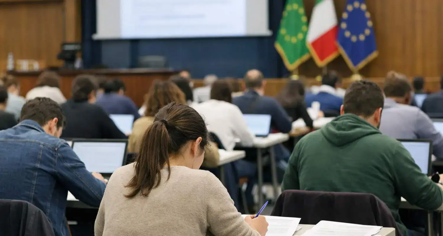 Alt text: Studenti e candidati durante un esame o prova di concorso pubblico in aula con bandiere italiane ed europee sullo sfondo.